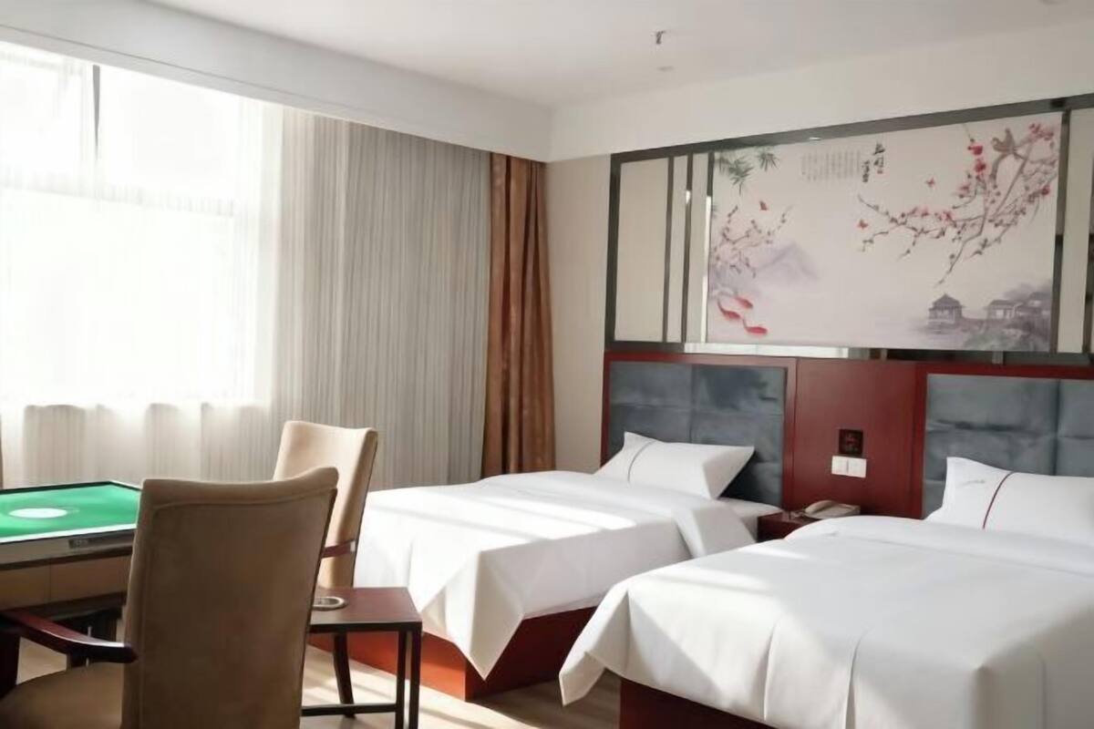 A hotel room with two beds, a desk with a green table, and a large painting on the wall.