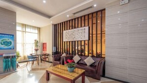 A hotel lobby with a checkered floor, a wooden coffee table with a flower vase, a brown leather sofa with cushions, a wall with vertical wooden slats, and a framed calligraphy piece. - Pinyi Hotel (Hefei Fanhua Avenue Zhonghuancheng) (Hefei)