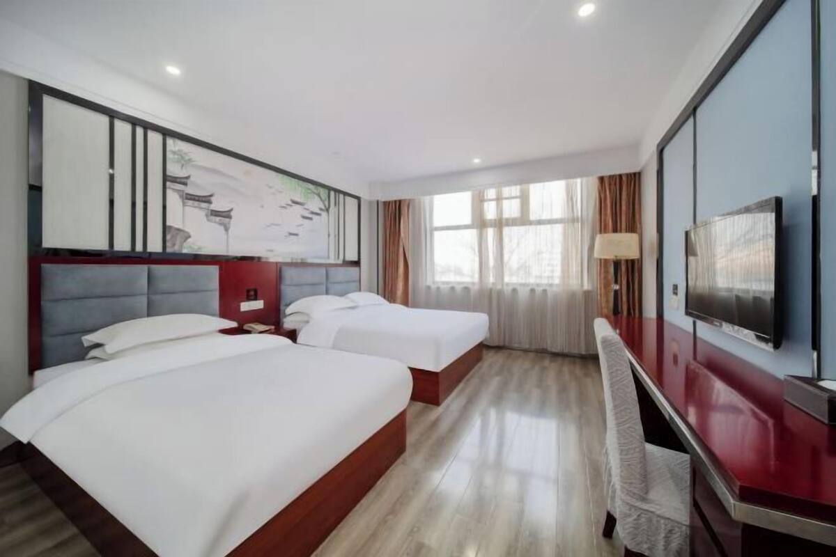 A hotel room with two beds, a desk, a TV, and a large window with curtains.