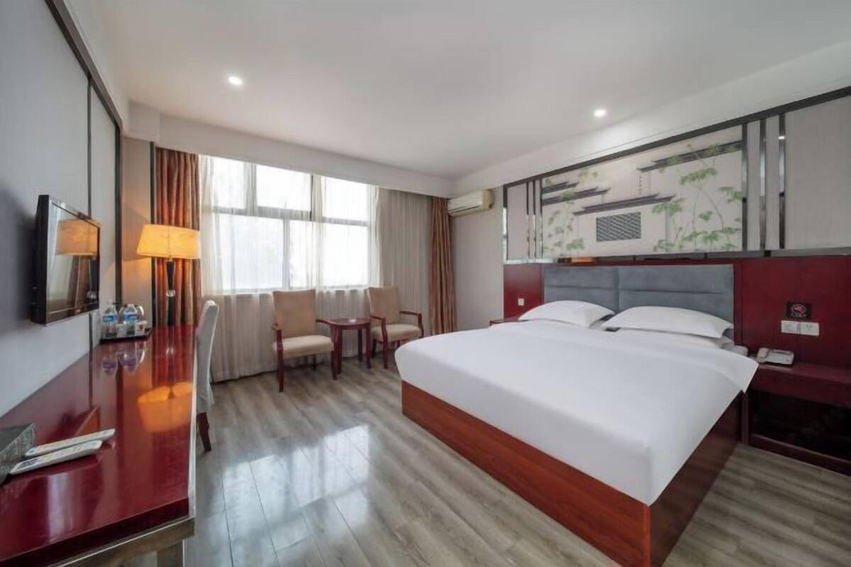 A hotel room with a large bed, a desk with a lamp, a chair, a TV, and a window with curtains.