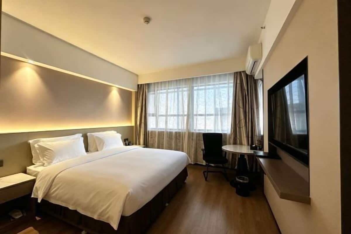 A hotel room with a bed, bedside table, chair, TV, and a window with curtains.