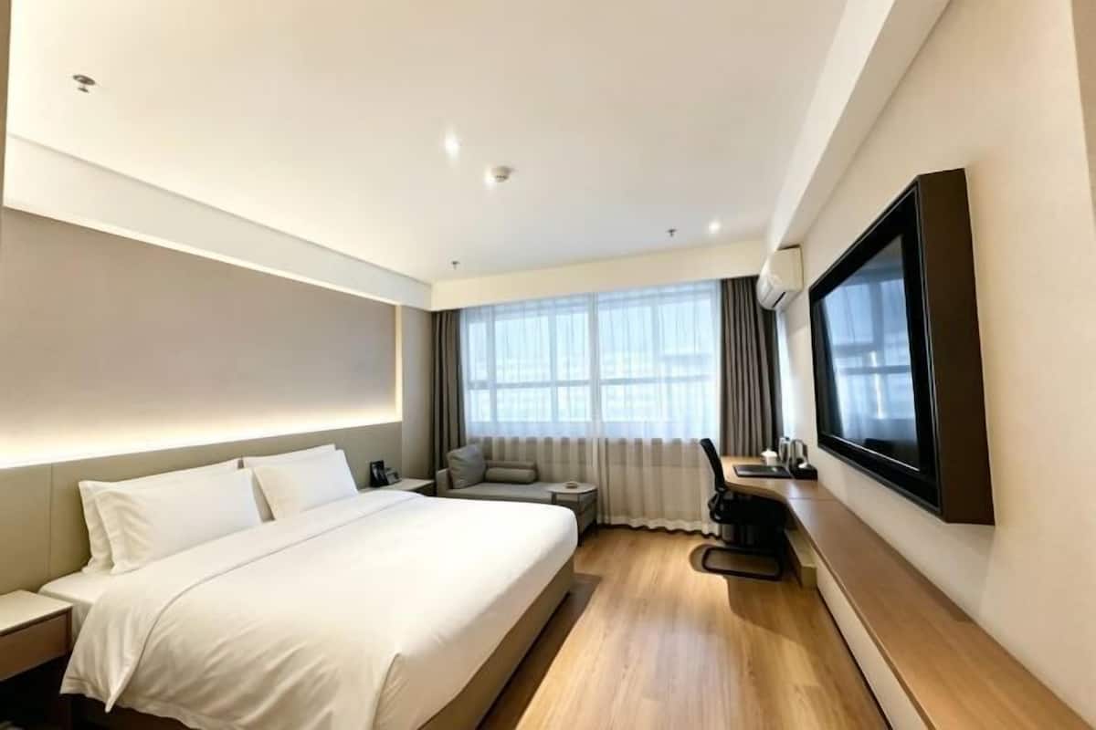 A modern hotel room with a large bed, a seating area, a flat-screen TV, and a large window.