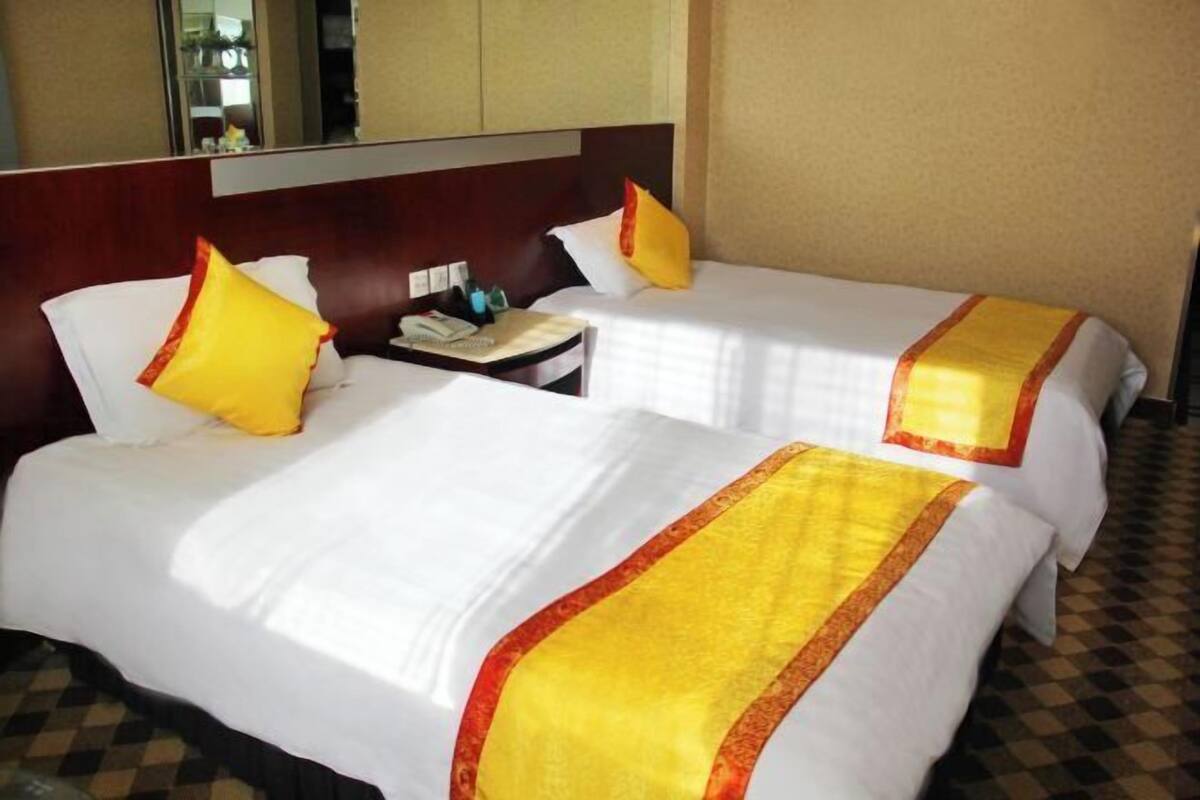 A hotel room with two beds, each with white linens and yellow accents, a bedside table with a phone and a lamp, and a checkered floor.
