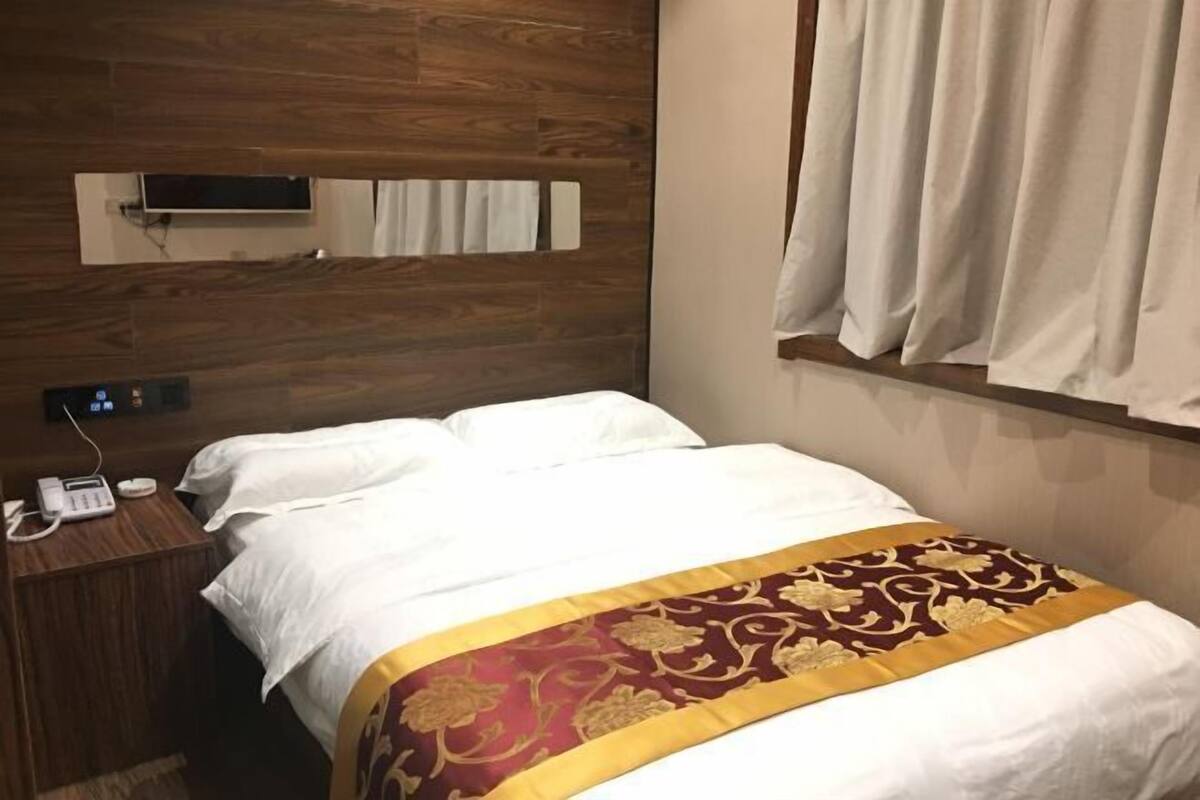 Ein Hotelzimmer mit einem hölzernen Kopfteil, einem Bett mit weißen Leinen und einer dekorativen Decke in Gold und Rot, einem Nachttischtelefon und einem Fenster mit Vorhängen.