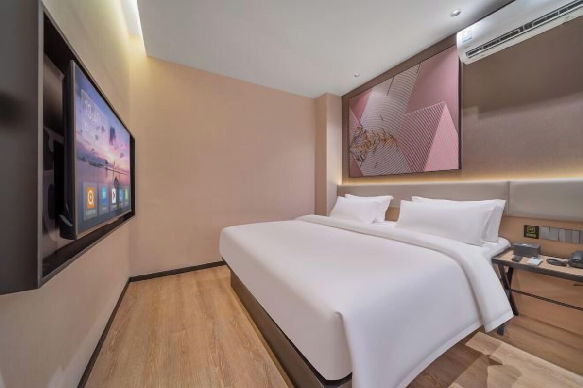 A modern hotel room with a large bed, a flat-screen TV mounted on the wall, a bedside table with a lamp, and a large abstract painting above the bed.