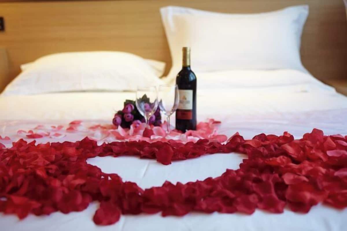 A bed with rose petals arranged in a heart shape, a bottle of wine, and a glass of wine on the bed.