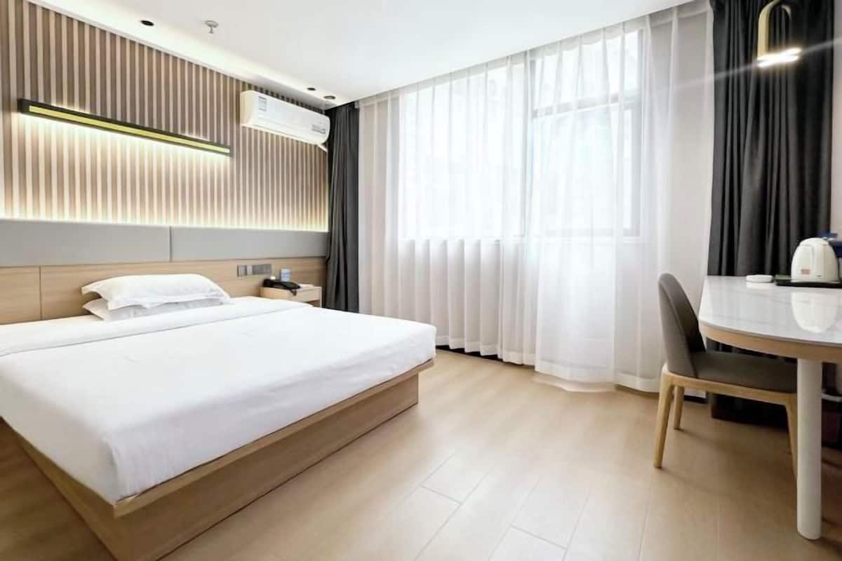A modern hotel room with a large bed, bedside table, and a dining area with a chair and table.