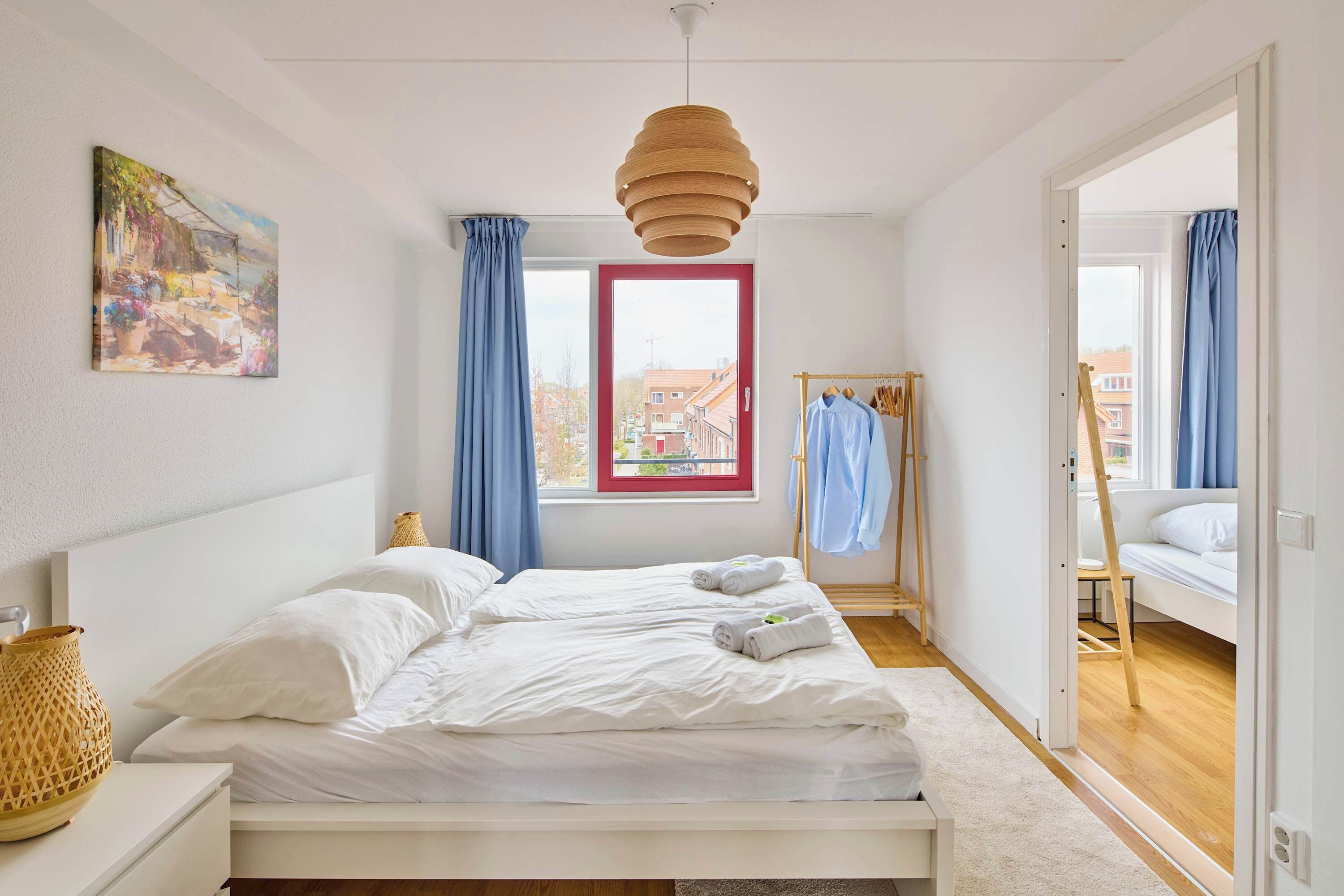 A bedroom with a bed, a window with curtains, a hanging light fixture, and a painting on the wall.