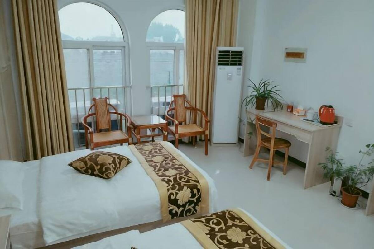 A hotel room with a bed, two chairs, a desk, a potted plant, and a view through large windows.