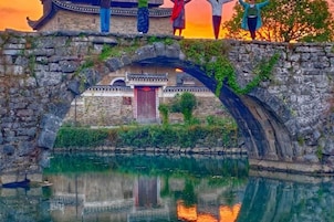 Un puente de piedra con un edificio tradicional al fondo, una masa de agua debajo y una puesta de sol en la distancia.