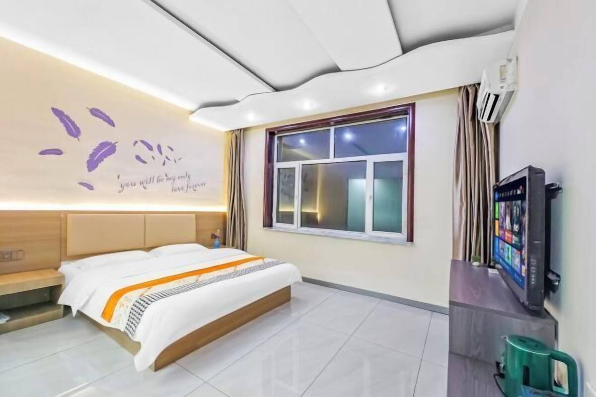 A modern hotel room with a bed, a television, a window, and a wall with a decorative phrase.