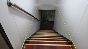 A staircase with wooden steps and a metal handrail, leading to a landing with a potted plant and a window with curtains. - Xiwuqi Jindi Hotel (West Ujimqin Banner)