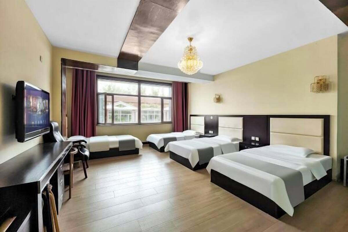A hotel room with a large bed, two smaller beds, a desk with a chair, a TV, and a window with curtains.
