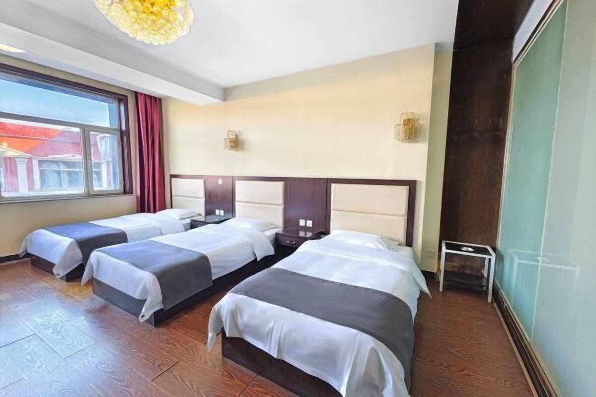 A hotel room with three beds, wooden flooring, a large window with curtains, and a modern chandelier.