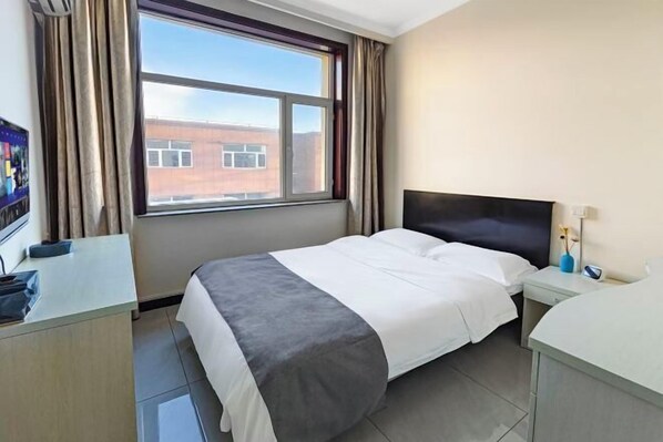 A hotel room with a bed, a desk, a nightstand, a window with curtains, and a view of a building outside.