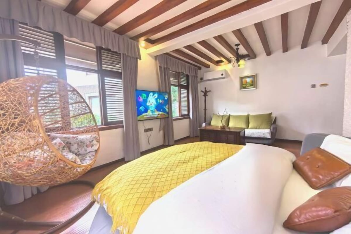 Un dormitorio con una cama grande, una silla colgante, un televisor y una ventana con cortinas.