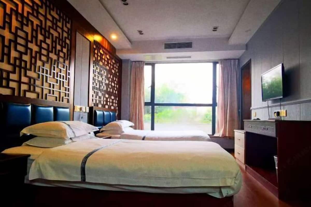 A hotel room with two beds, a TV, a desk, and a large window with curtains.