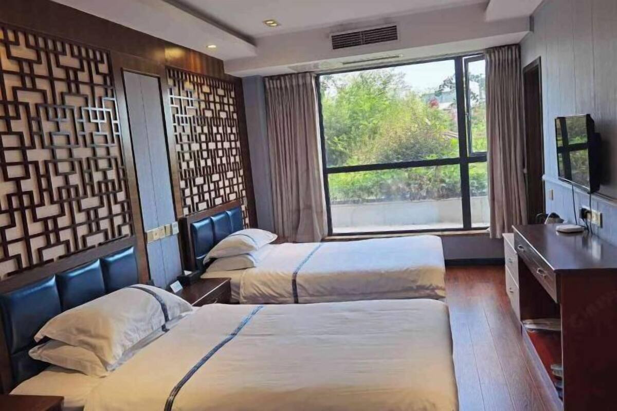 A hotel room with two beds, a TV, a desk, and a large window with a view of greenery.