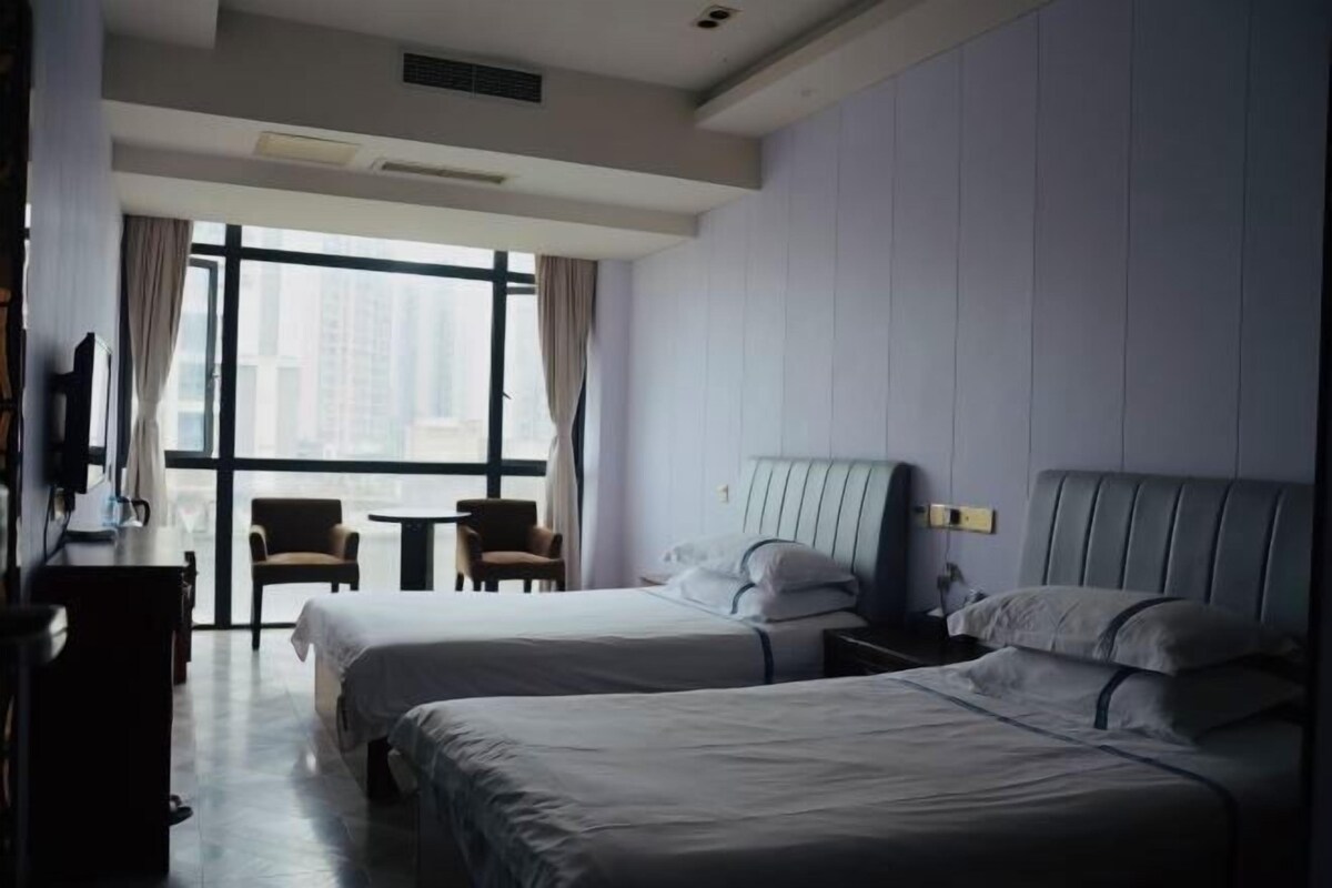 A hotel room with two beds, a desk, a chair, and a large window offering a city view.
