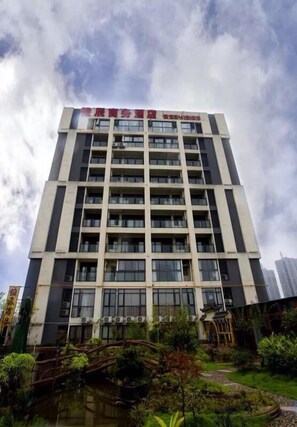 A multi-story building with a flat facade, numerous windows, and a landscaped area with a bridge and pond. - Junchen Business Hotel (Chongqing)