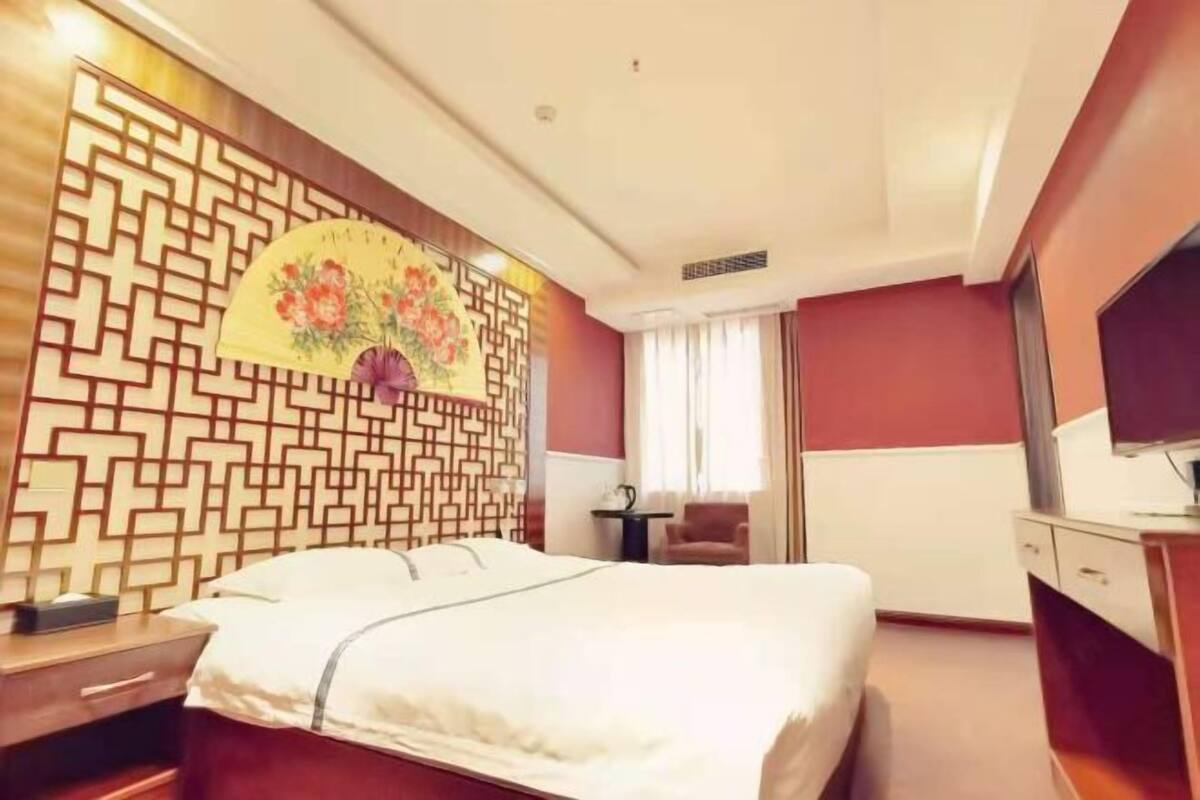 A hotel room with a bed, a desk, a chair, a TV, and a decorative wall with a fan.