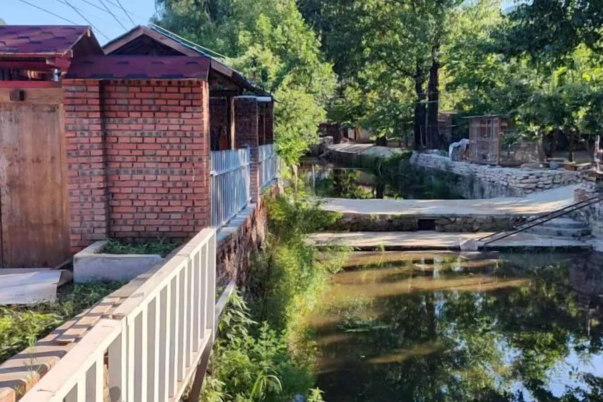 Un bâtiment en briques surmonté d’un toit rouge, avec une clôture en bois et un étang traversé par un pont de pierre.