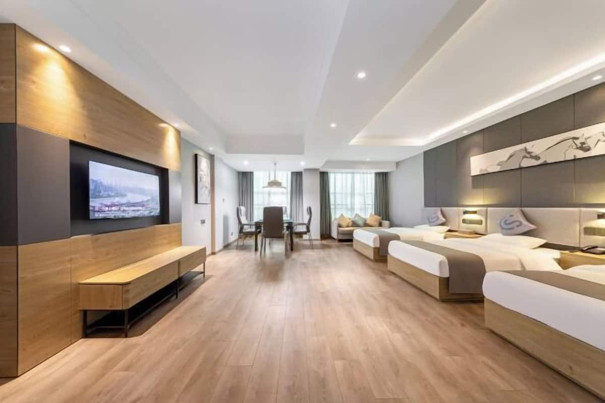 A modern hotel room with a wooden floor, a TV mounted on the wall, a dining area with a table and chairs, and a bed with a headboard.