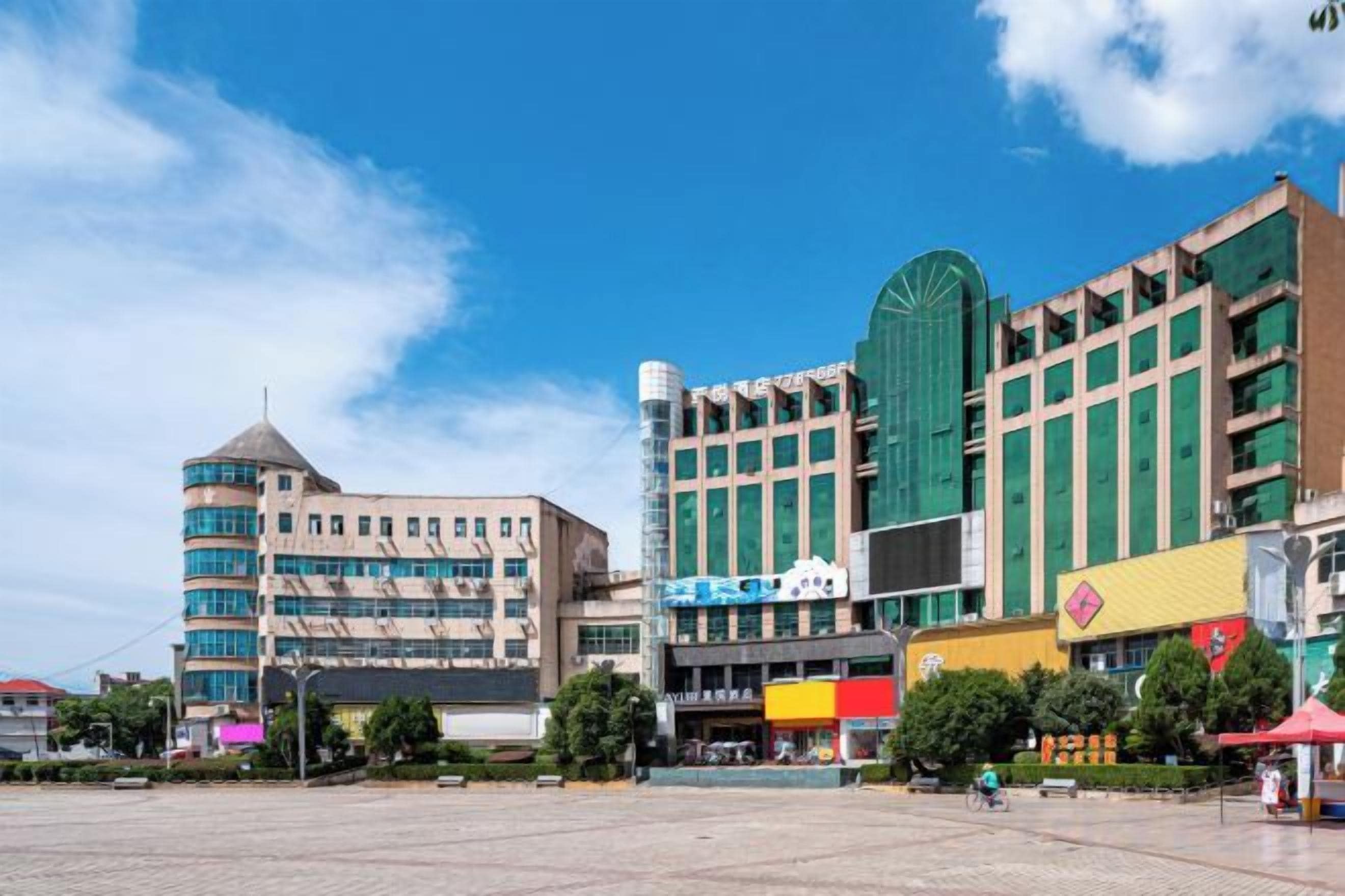 A modern urban square with a variety of buildings, including a prominent green structure with a dome and a beige building with a rounded corner.