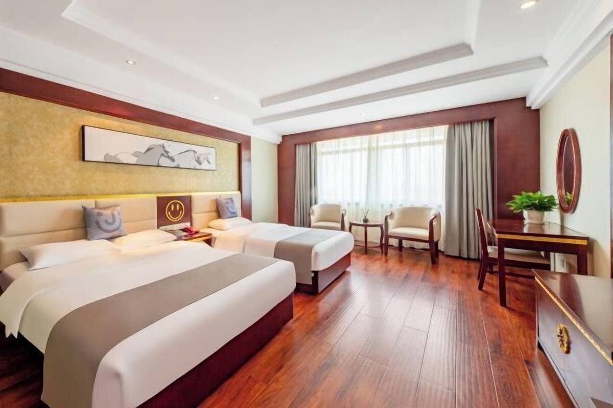 A hotel room with two beds, a desk, a chair, a small table, and a large window with curtains.