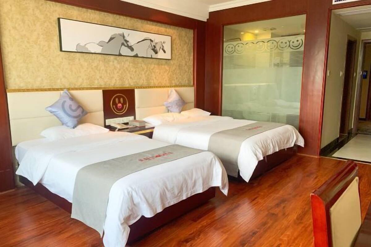 A hotel room with two beds, a wooden floor, a framed artwork on the wall, and a glass door leading to a bathroom.