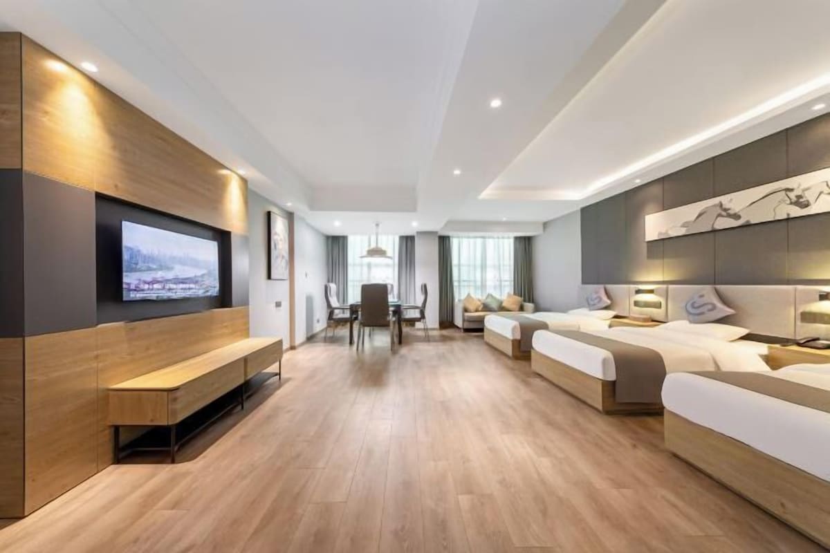 A modern hotel room with a wooden floor, a large flat-screen TV mounted on the wall, a sofa, a bed, and a dining area with a table and chairs.