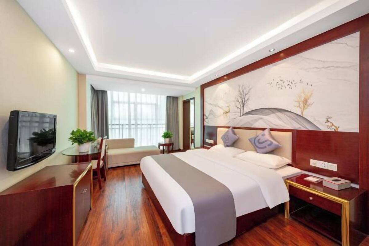 A modern hotel room with a large bed, a flat-screen TV, a dining area with a table and chairs, and a large window with curtains.