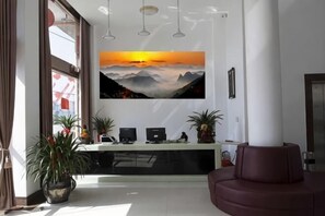 A reception area with a desk, computers, potted plants, and a large painting of a sunset over mountains. - Gorgeous Hotel (Yueqing)