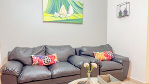 A living room with a dark leather sofa, colorful pillows, a vase with flowers, and a painting on the wall. - Garden Condos Apt 44 - Comfy 1br Apt In Sosua! (Sosúa)