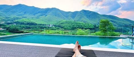 Una persona descansando en una silla de descanso junto a una piscina, con vistas a montañas y vegetación.