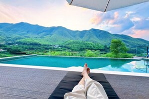 A person relaxing on a lounge chair by a pool with a view of mountains and greenery.
