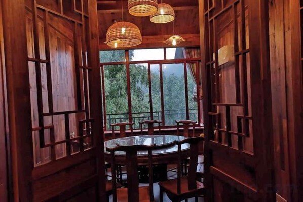 A wooden dining area with a round table and chairs, large windows, and hanging light fixtures.