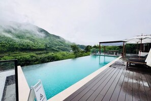 Une piscine à débordement avec une terrasse en bois, offrant une vue imprenable sur une vallée verdoyante et une montagne.