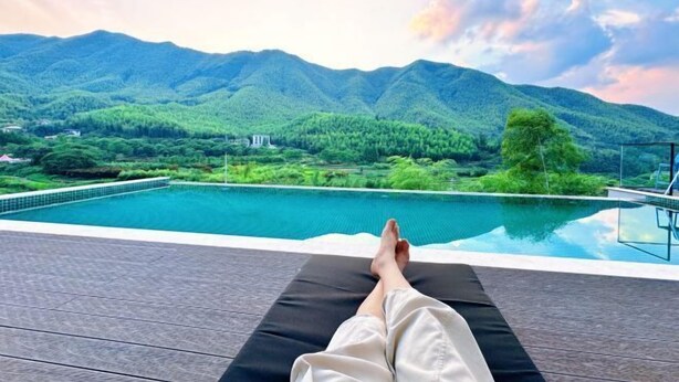 Une personne se détend sur une chaise longue au bord d’une piscine, avec une vue sur les montagnes.