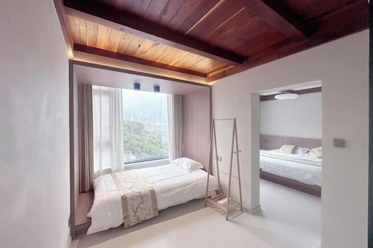 A bedroom with a large window, a bed, a wardrobe, and a ceiling with exposed beams.