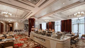 A hotel dining area with a buffet setup, a large carpeted floor, and a chandelier.