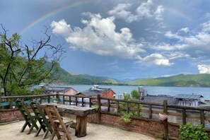 Un patio con una mesa y sillas, con vista a un lago y edificios, y montañas al fondo.