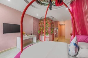 Ein Schlafzimmer mit einem Bett, einem Fernseher, einer weißen Kommode, einer rundförmigen Pflanzenstruktur und einem rosa Vorhang.