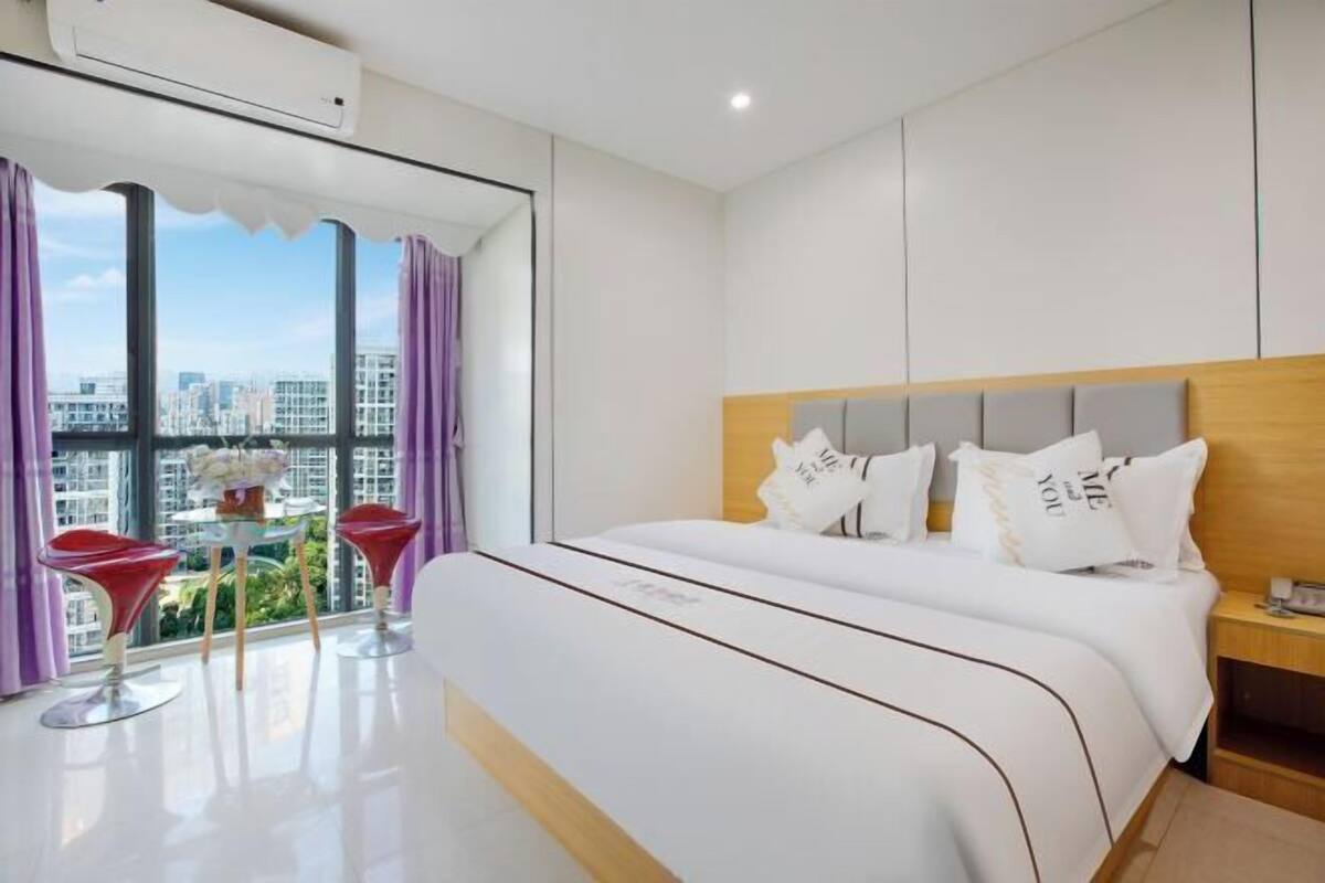 Een moderne hotelkamer met een groot bed, een klein tafeltje met stoelen en uitzicht op de stad door grote ramen.