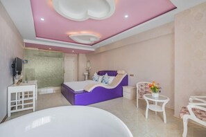 Ein modernes Schlafzimmer mit einem violetten Bett, einer weißen Badewanne und einem kleinen runden Tisch mit Blumen.