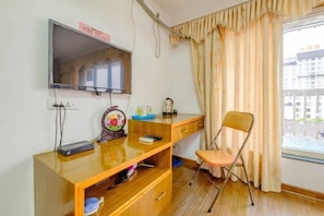 Quarto com escrivaninha de madeira, televisão na parede, uma cadeira e janela com cortinas.