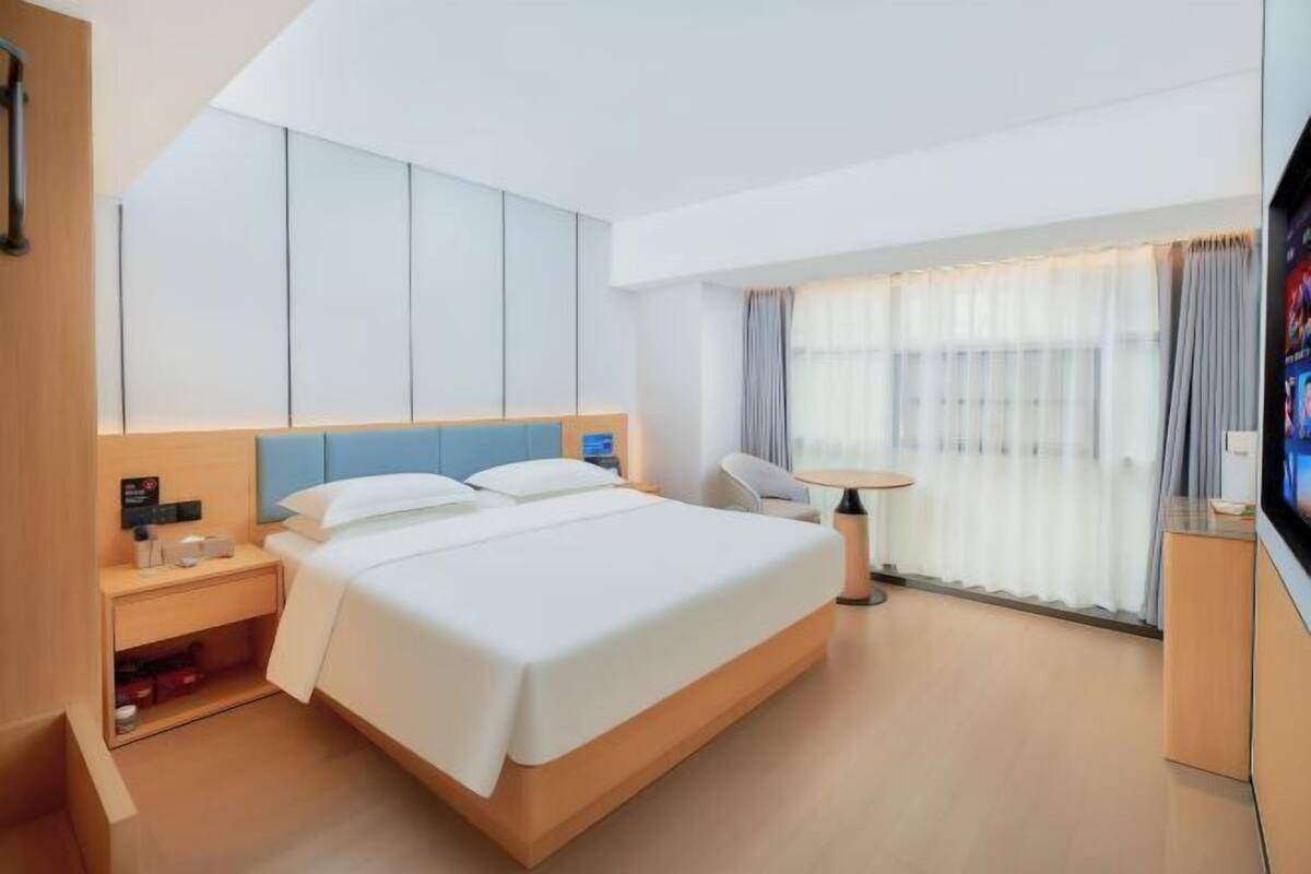 A modern hotel room with a large bed, a bedside table, a small desk, a chair, and a TV.
