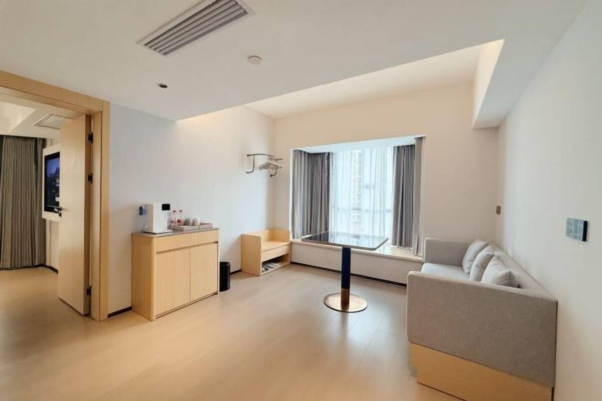 A modern hotel room with a sofa, a small table, a desk with a chair, a window with curtains, and a door leading to another room.