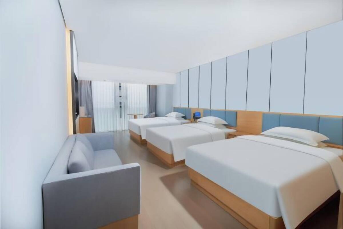 A hotel room with three beds arranged in a row, a sofa, and a large window with curtains.