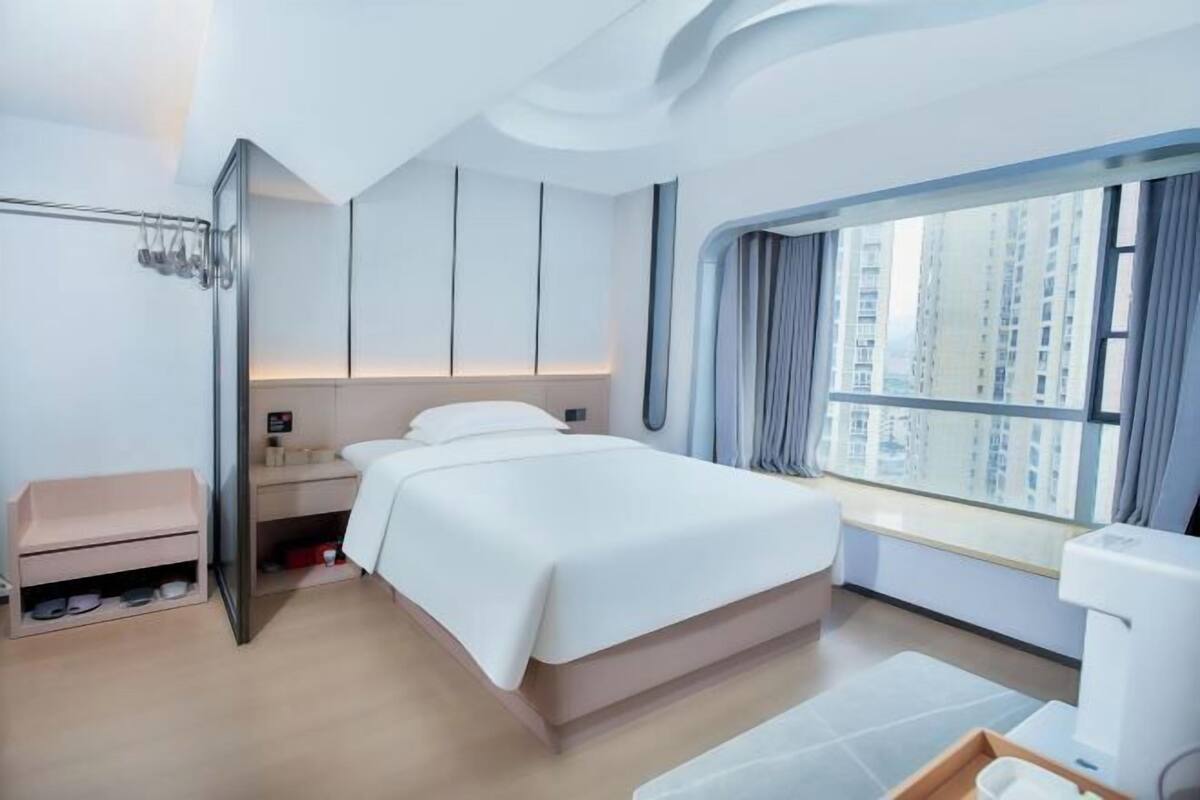 A modern hotel room with a large bed, a built-in wardrobe, a vanity with a mirror, and a window with a city view.